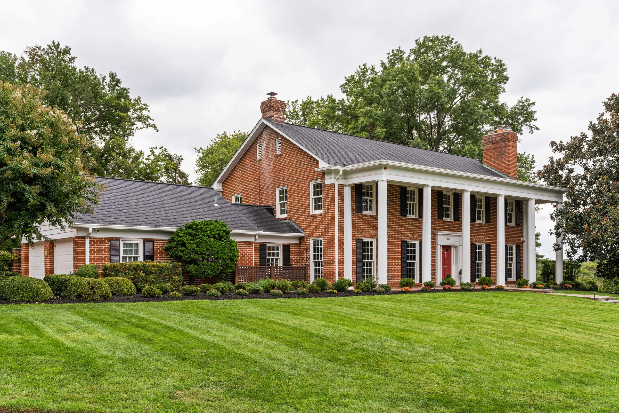Front of a brick colonial home with white columns and a wide green lawn under a flat, overcast grey sky