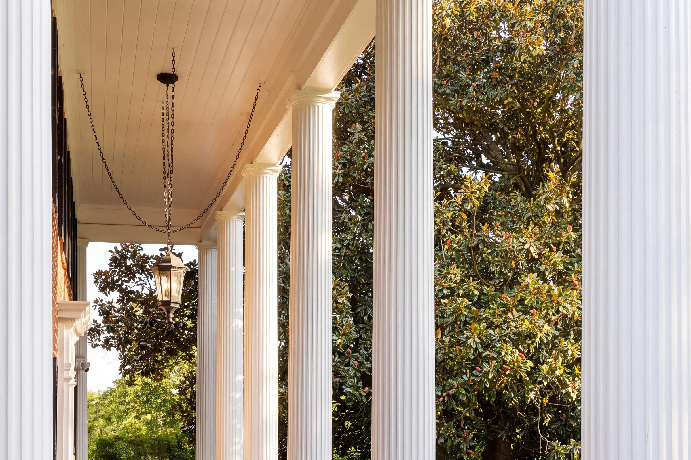 After: the same porch in AI Studio with warm sunlight on the columns and magnolia leaves, every flute and chain link intact